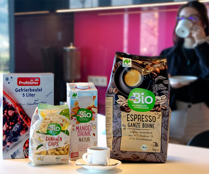 Neue Partnerschaft: dm-Produkte jetzt auch für Ihr Büro Produkte von dm stehen auf einem Tisch. Im Hintergrund trinkt eine Frau Kaffee.