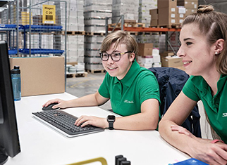 Ergonomie in Produktion und Logistik – Wohlbefinden und Effizienz am Arbeitsplatz Zwei Mitarbeiterinnen im Lager der Streit Service & Solution GmbH & Co. KG arbeiten an einem ergonomisch höhenverstellbaren Schreibtisch.