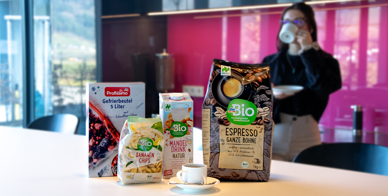 Die dm-Eigenmarken Produkte von dem stehen auf einem Tisch. Im Hintergrund trinkt eine Frau Kaffee
